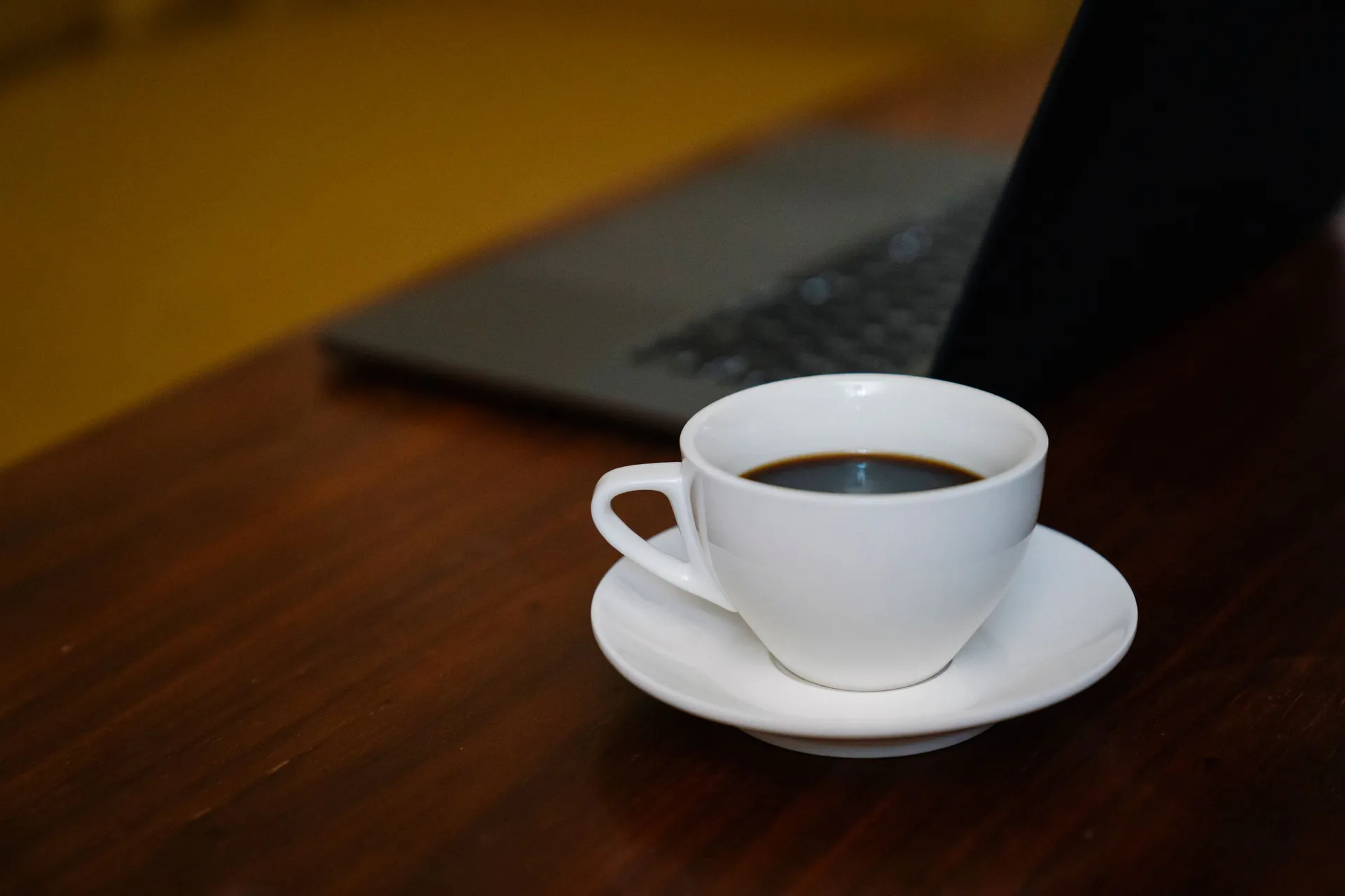 Cup of black coffee on a saucer beside a laptop on a wooden desk in warm light